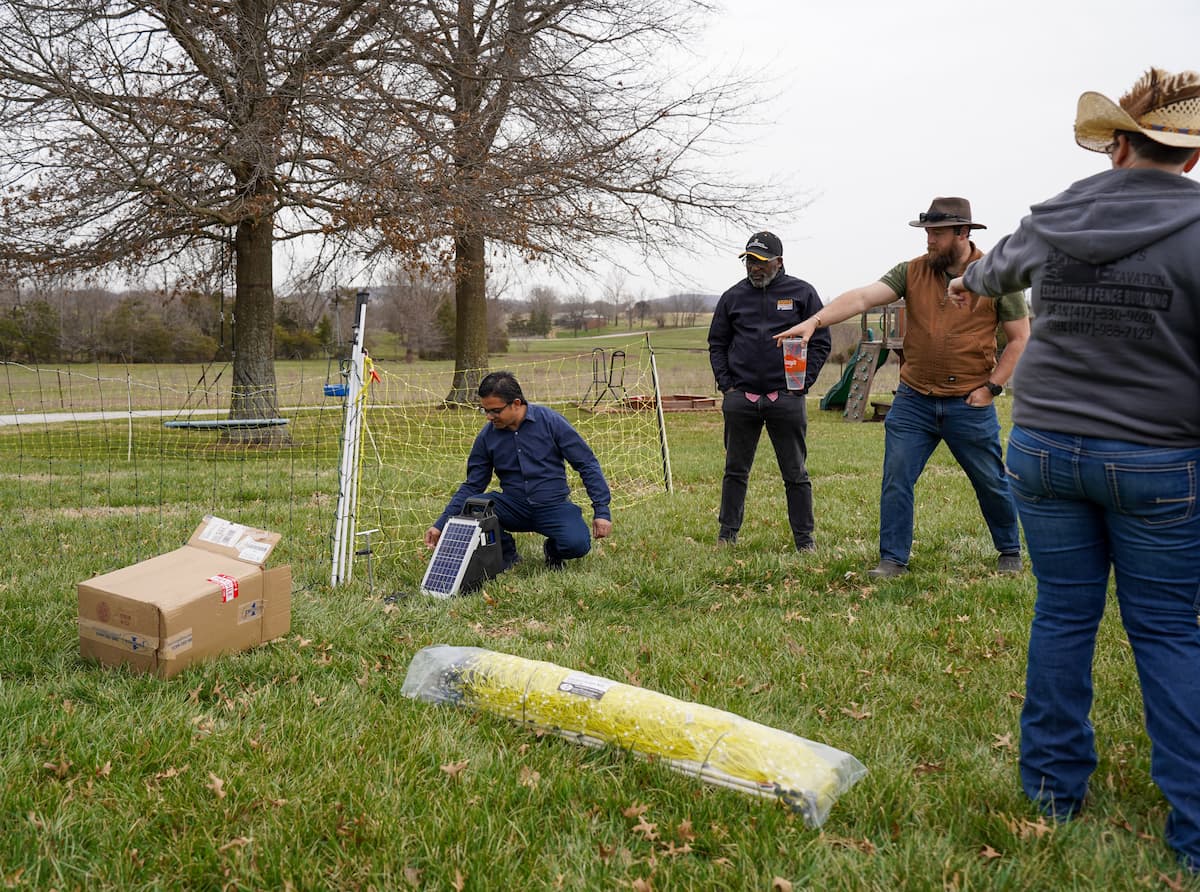 A standing man points at another man bent down beside a fence.