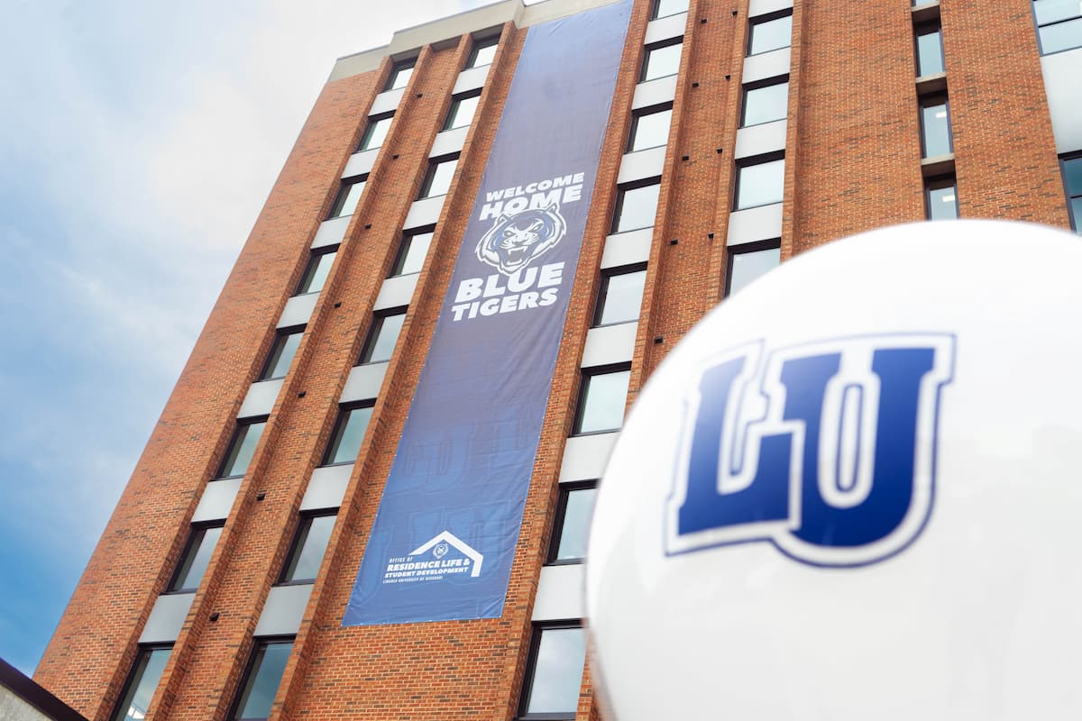 Large “Welcome Home Blue Tigers” banner hangs on tall brick building, with LU logo balloon in foreground under blue sky.