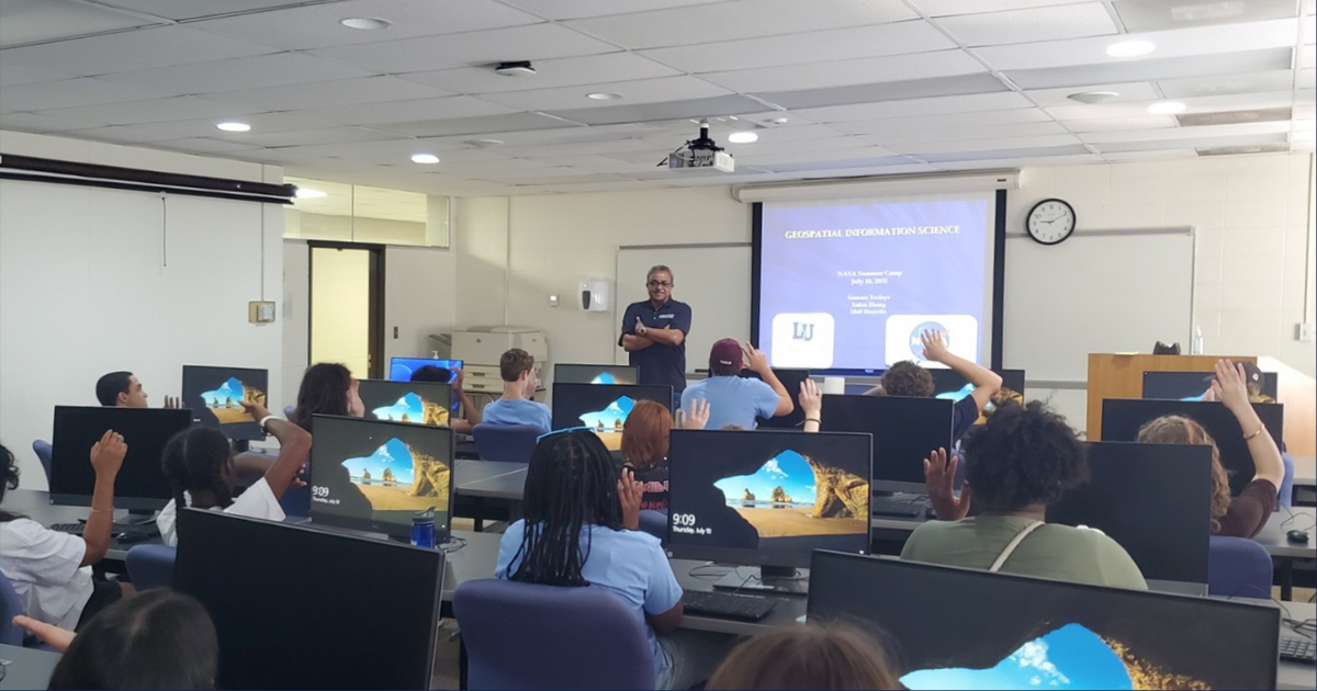 A group of students sit behind computers in a classroom, watching a presentation.
