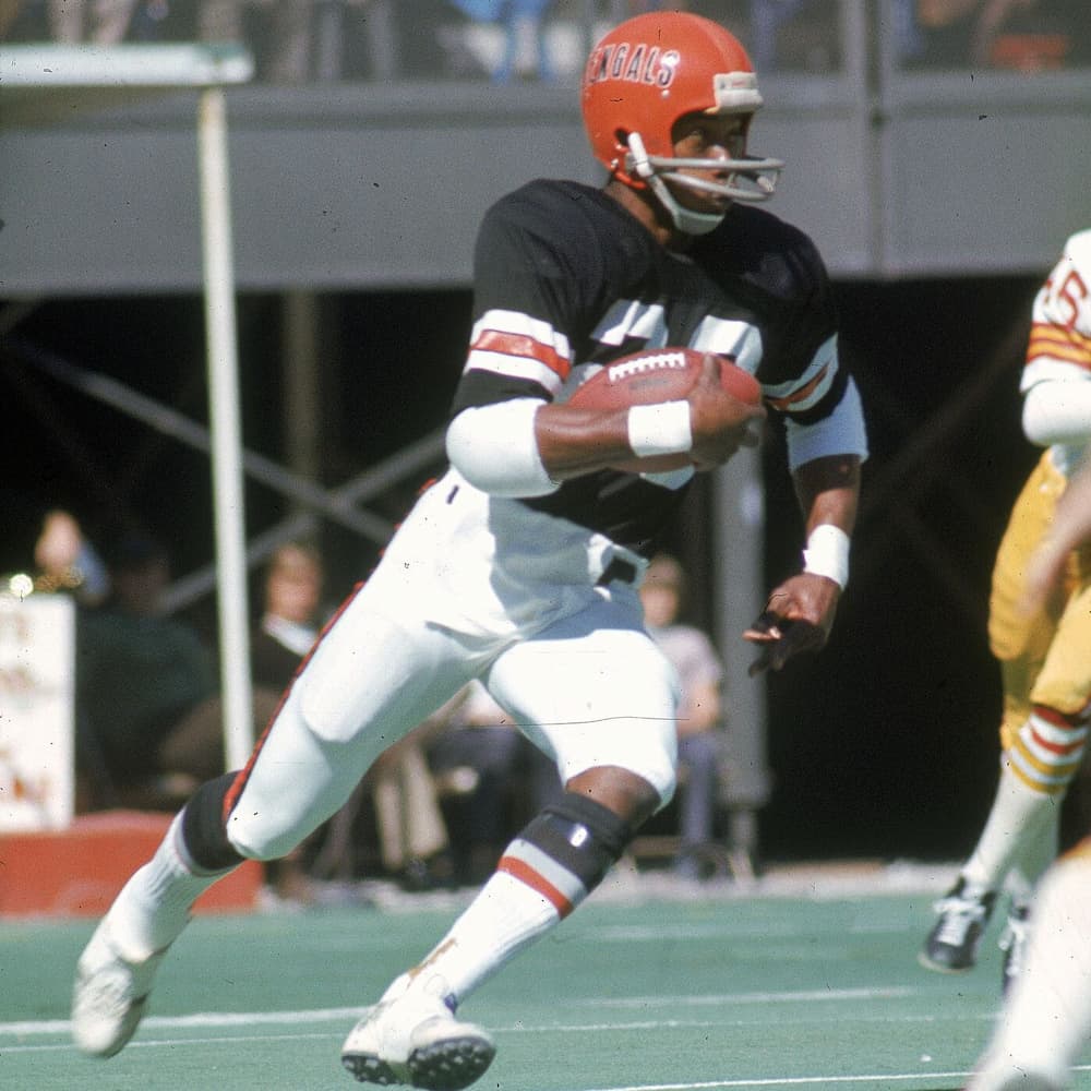 Football player wearing a Cincinnati Bengals uniform, including a black jersey with white and red stripes and a red helmet with “BENGALS” lettering, running with the ball tucked under his arm during a game.