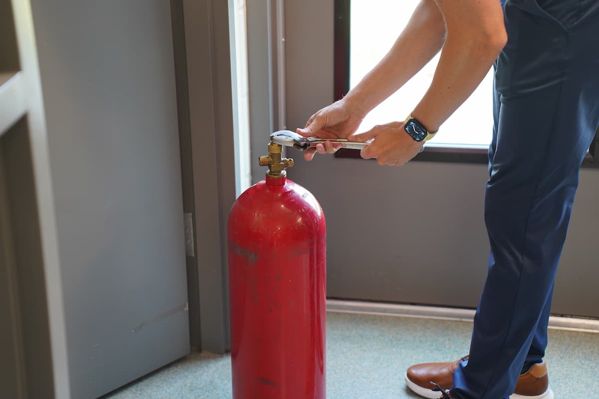 A man unscrews a valve on a red canister using a wrench.