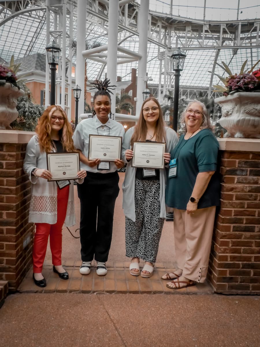 Three young woman holding certificates stand next to an older woman.