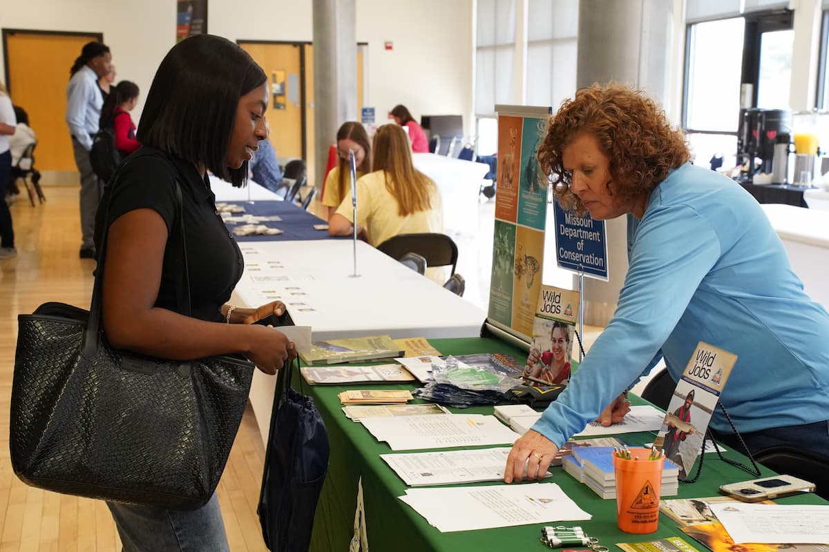 A woman behind a table points to a stack of papers a person on other side of table is looking at.