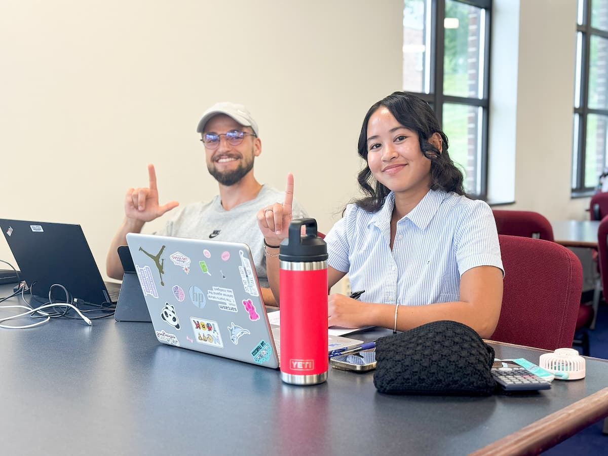 A male and a female student sit at a table while working on their laptops and smiling at the camera.