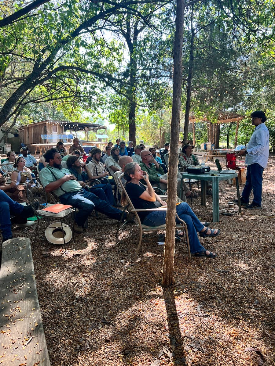 A man stands in front of group of people sitting around a table outdoors.