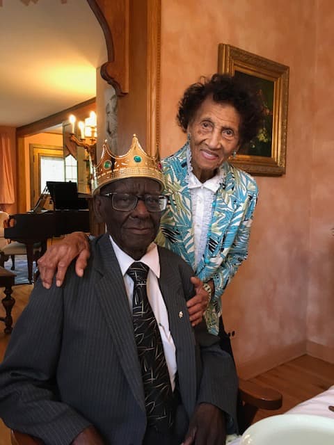 Edward Harrison sits at a table while his wife, Frances, has her hand on his shoulders and smiles for a photo.