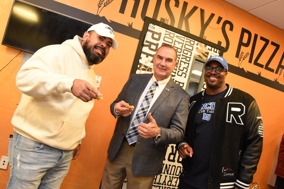 Three men smile at the camera while holding pizza at the Lincoln University of Missouri Slim & Husky's grand opening.