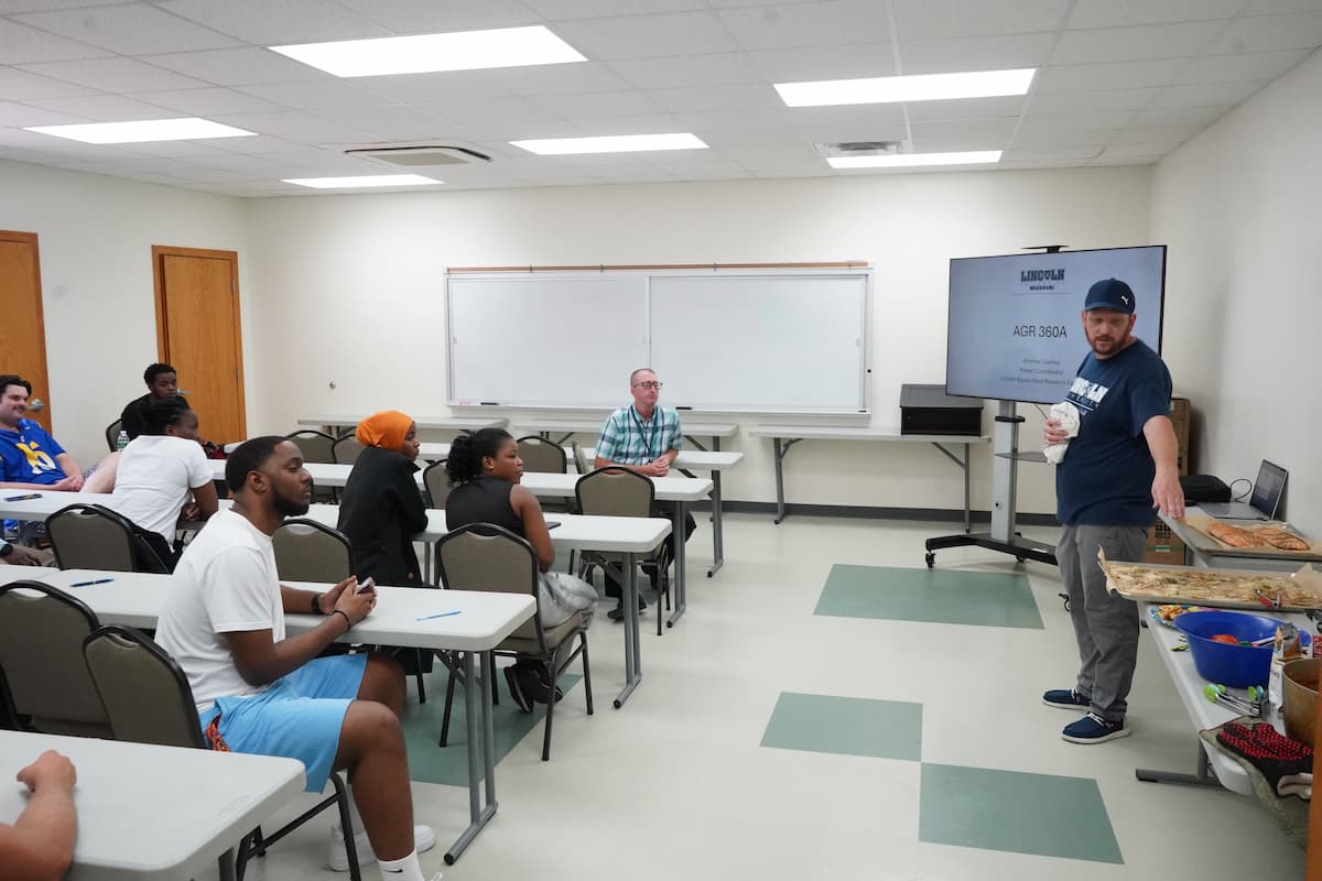 A man pointing at items on a table in front of a classroom of people.
