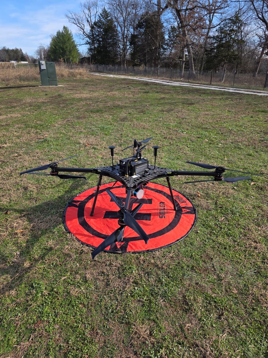 A large, black drone with eight rotors sits on a round, red landing pad in the grass.