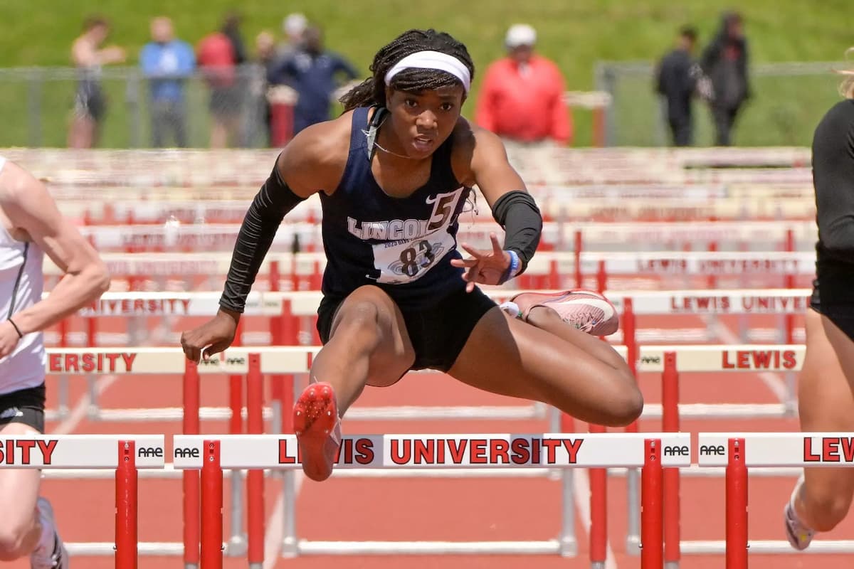 A female LU student jumps over a hurdle in a blue track uniform.