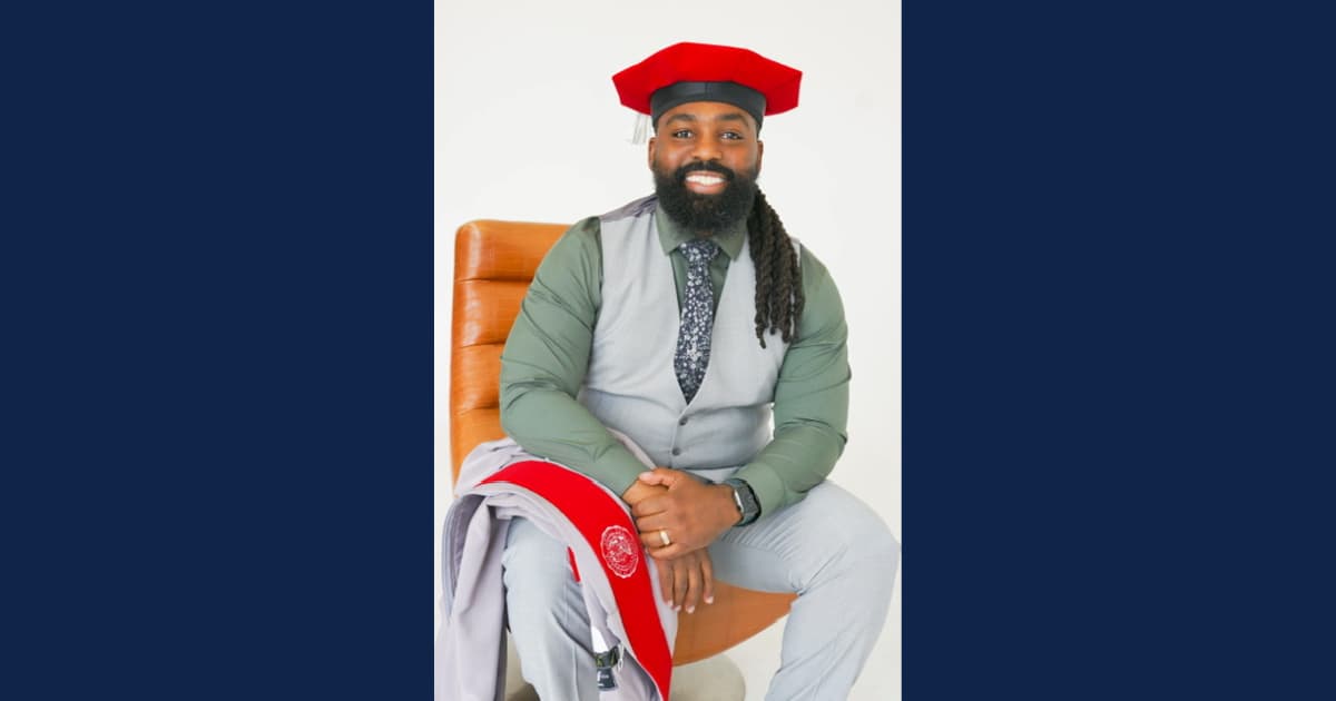 Joseph Simmons Jr. poses in doctoral regalia with a gray gown, red accents, and a red doctoral cap, marking his achievement as a Ph.D. graduate in special education.