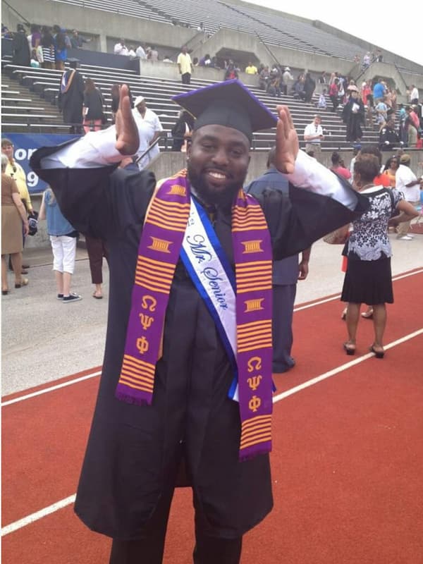Joseph Simmons Jr. smiles while celebrating graduation at Lincoln University, wearing a black cap and gown with an Omega Psi Phi stole and honor sash on a track field with stadium seating in the background.