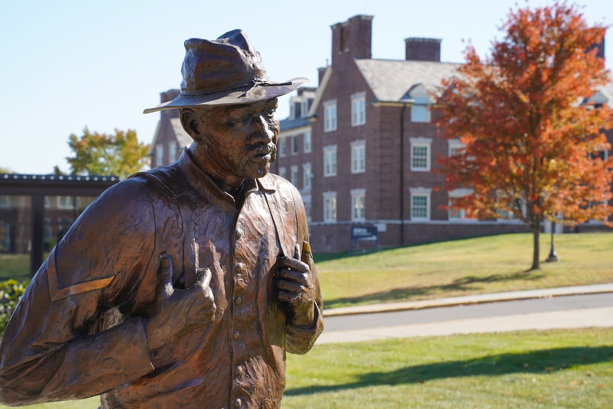 A bronze statue of a soldier stands with a tree and brick building in the background.