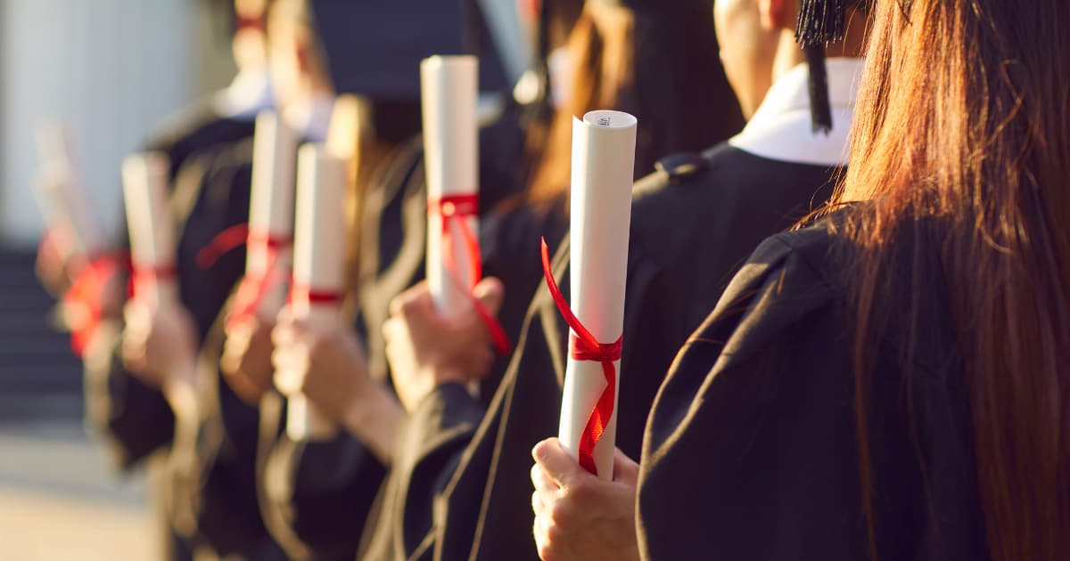 Graduate students in caps and gowns holding rolled diplomas tied with red ribbons during a commencement ceremony.