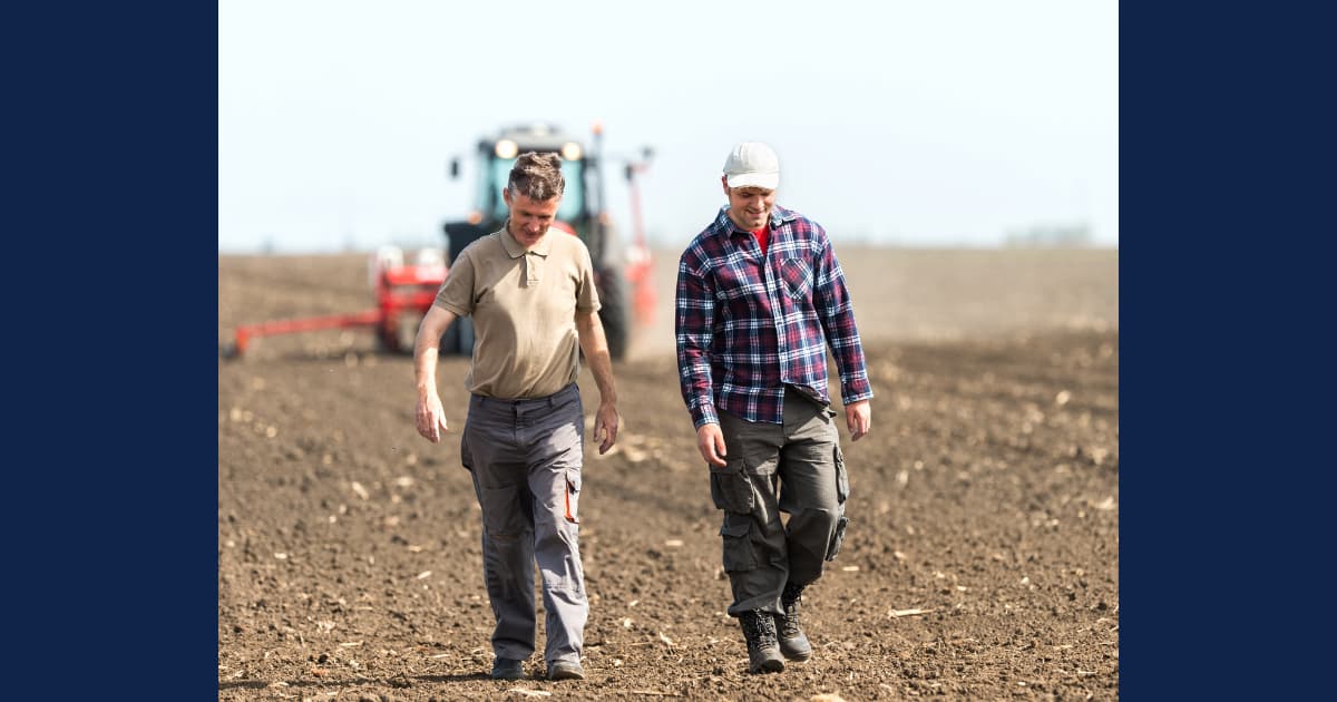 Two agricultural professionals walking through a cultivated field with a tractor in the background, representing sustainable farming and modern agricultural practices.