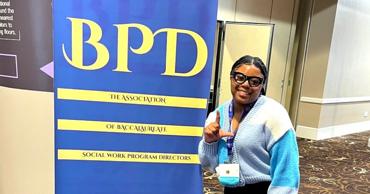 A young female students stands in front of a blue sign while making an L with her hands.