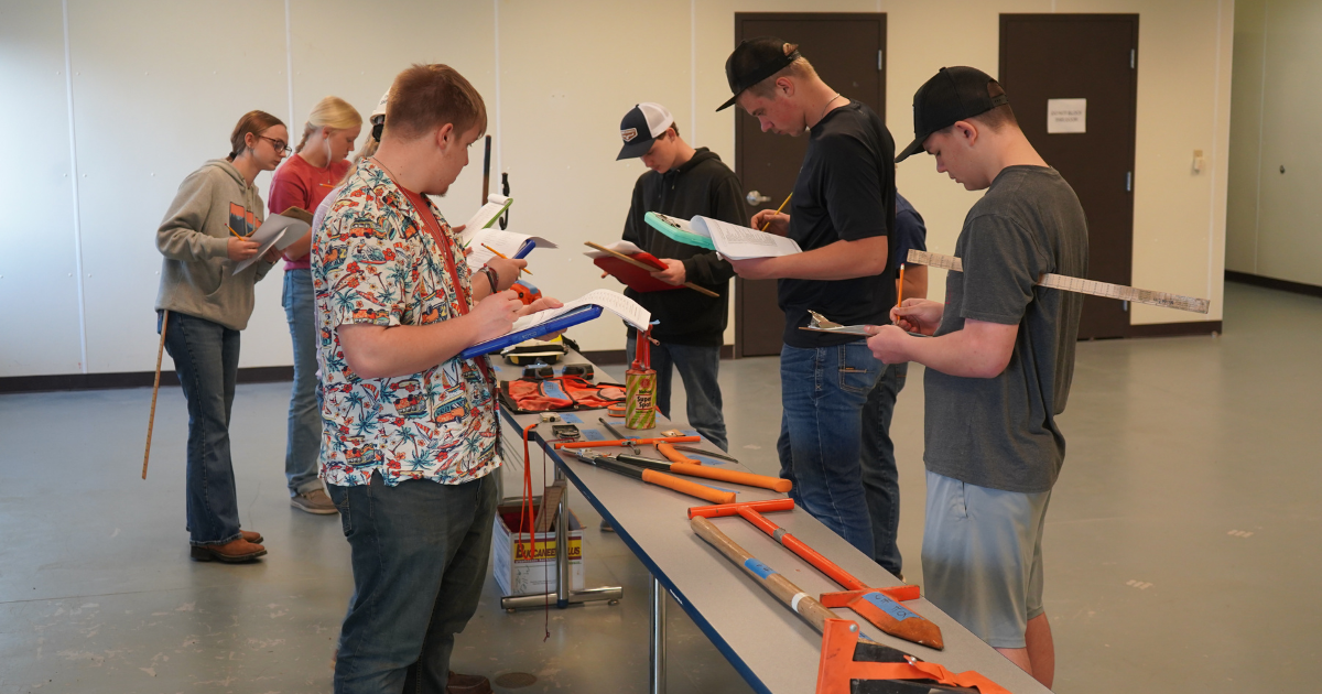 Six people standing around a table with various tools, including saws and clamps, all holding clipboards.