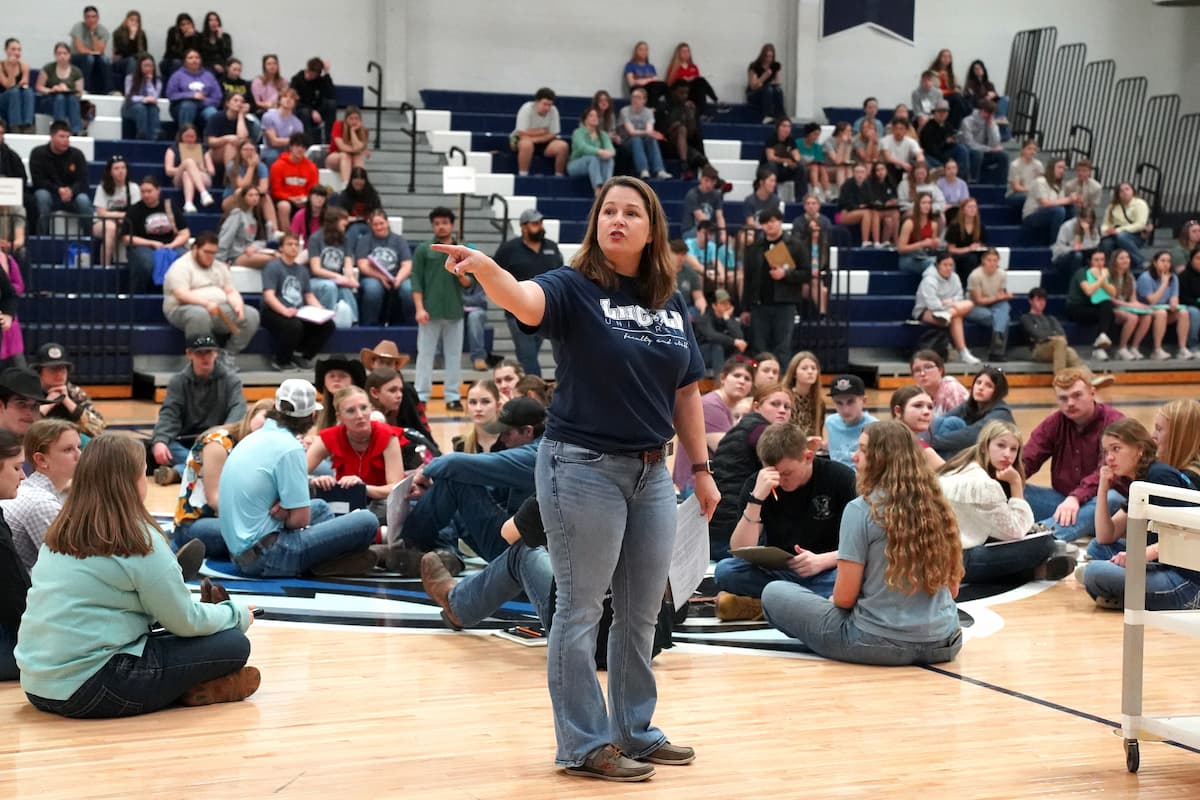 A gymnasium filled with students seated on bleachers and floor, with one person in the center of the floor pointing. 