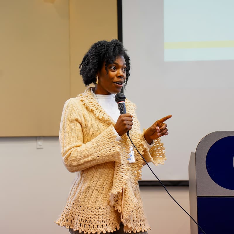 A woman stands speaking with a microphone in hand.