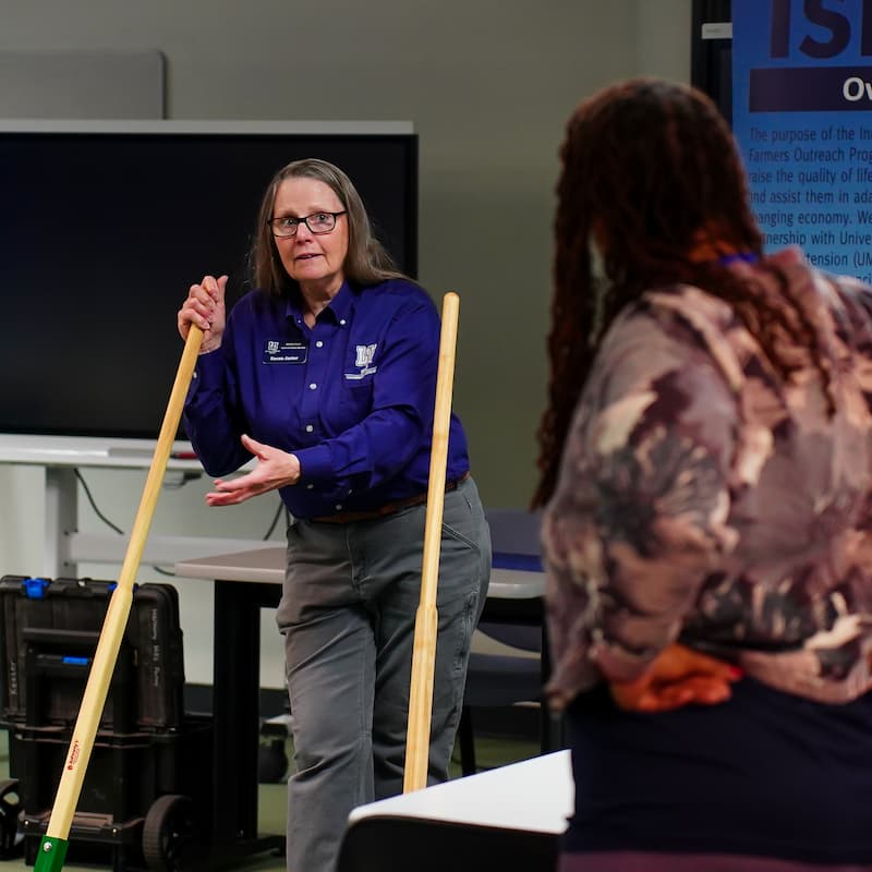 A woman holding a long-handled farm tool speaks to another woman.