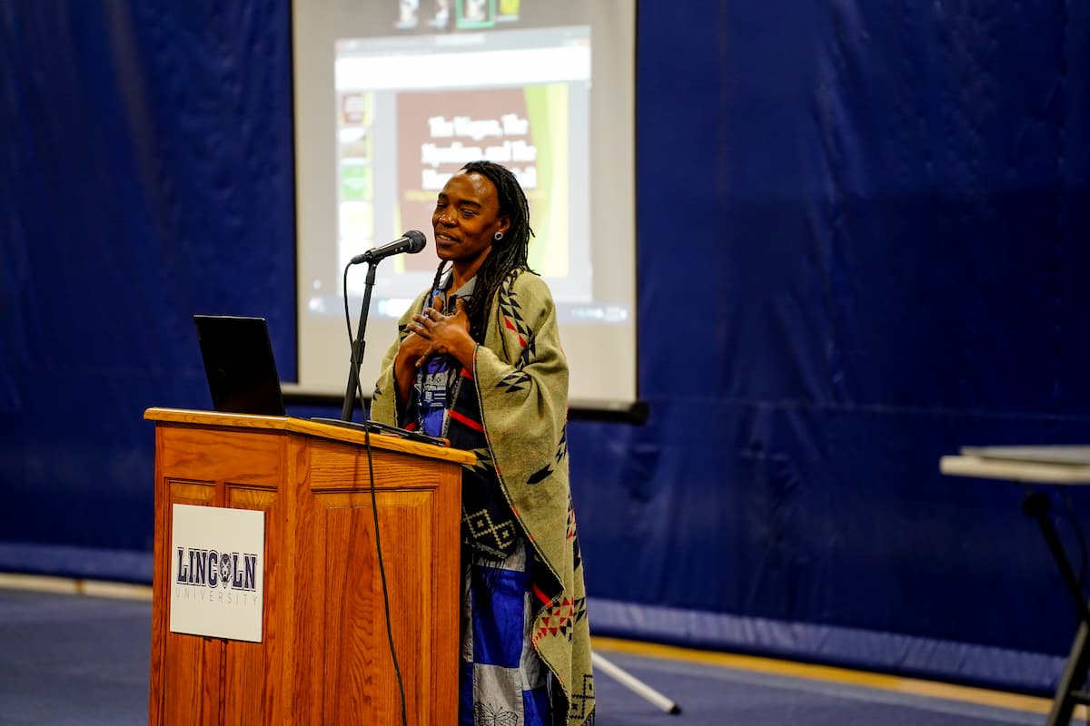 A woman stands at a lectern with her eyes closed and hands folded across her chest.