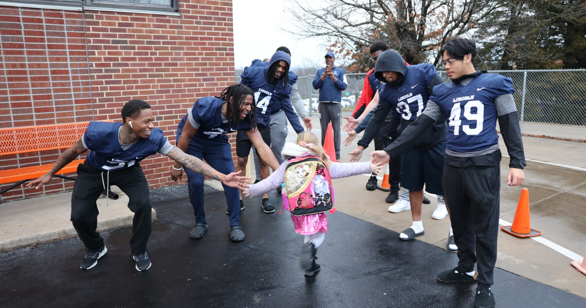 A group of Lincoln University football players in blue jerseys high five an elementary student as they walk into school with their backpack.