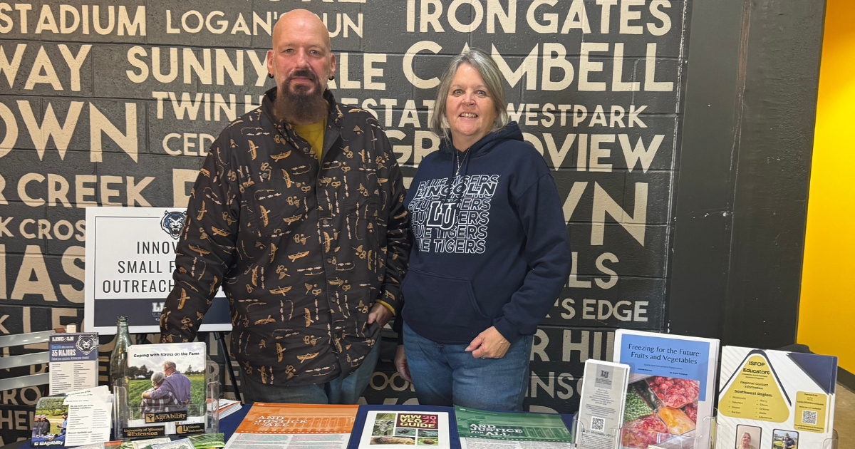 A man and woman stand behind a table covered with flyers, factsheets, and other paper handouts. 