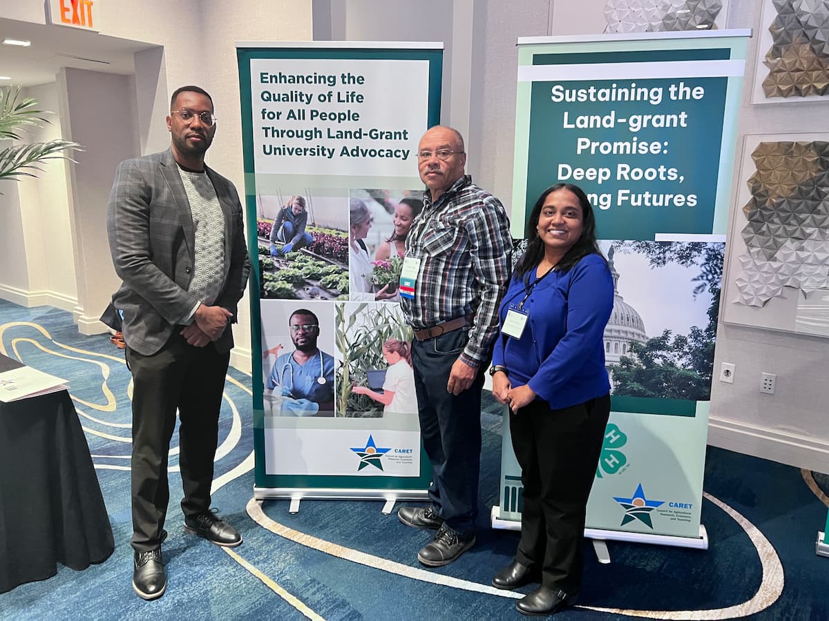 Three people standing in front of two vertical banners promoting Land-Grant University advocacy and sustainability.