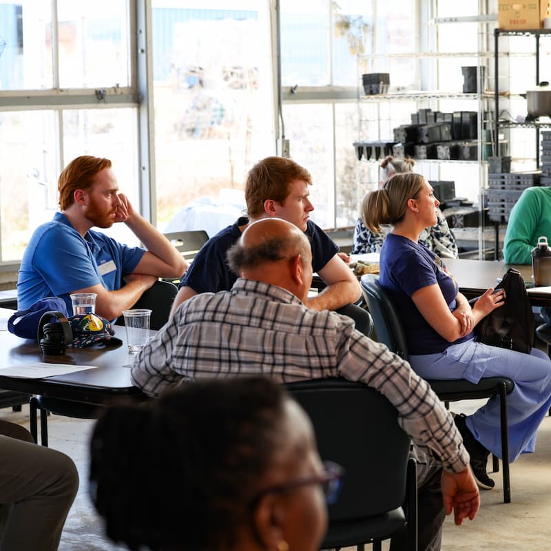 People sit at tables inside a greenhouse classroom, listening during a composting workshop.