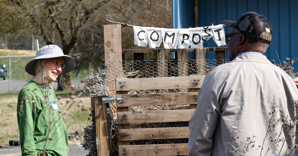 Lea Langdon talks with a participant beside a wooden compost bin labeled “COMPOST” during an outdoor workshop.