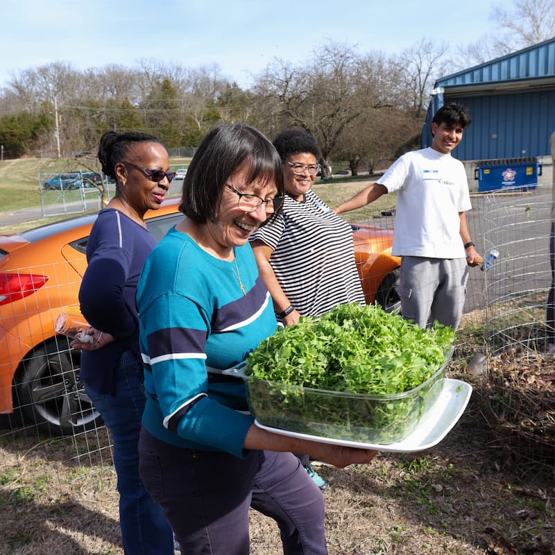 Workshop participants stand outside as Nadia Navarrete-Tindall brings greens to add to a compost pile in progress.