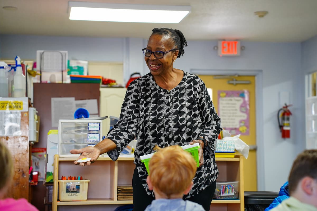 Woman stands in a classroom and engages students. She holds a plastic bin in one hand and a toy fried egg in the other. 
