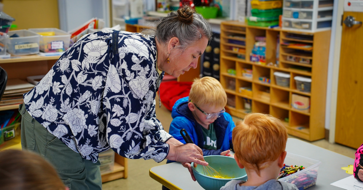 Woman leans over a table with children seated at it. She is holding a large bowl.