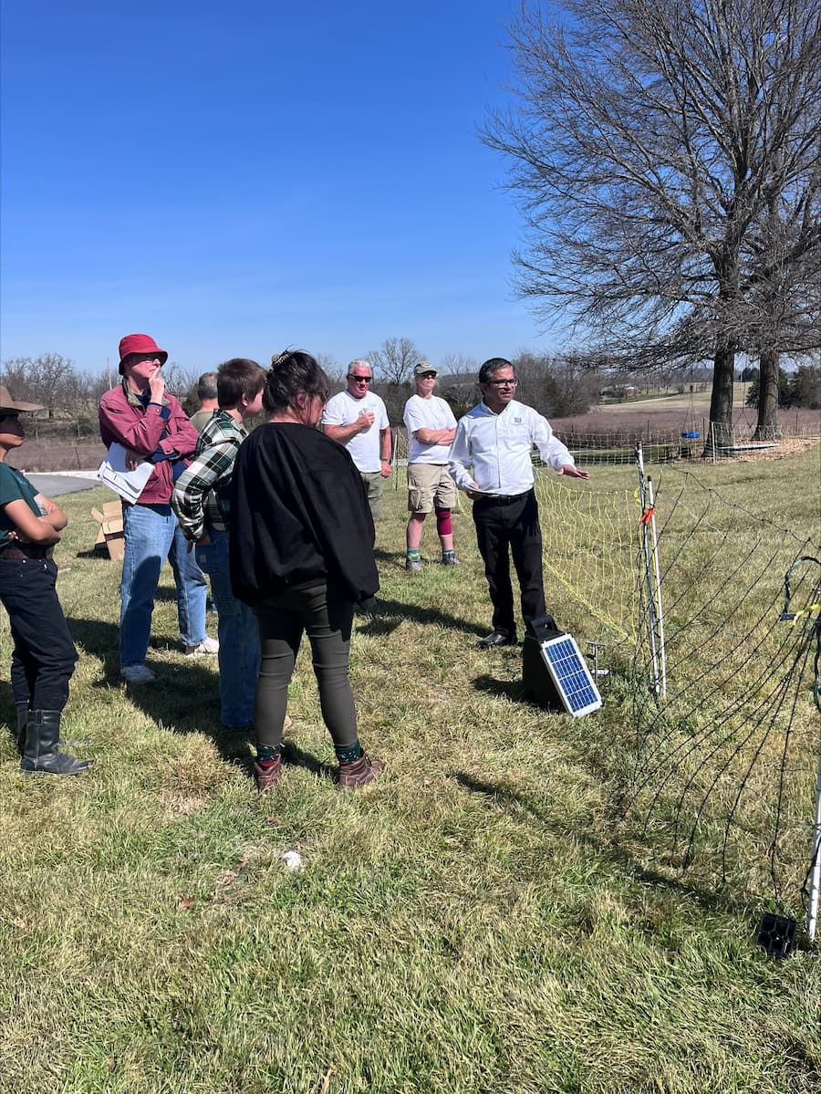A group of people gathered outdoors around a man explaining an electric fence setup.