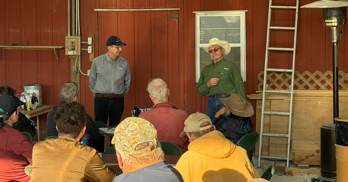 A group of people seated and listening to two men standing and speaking in front of a red wooden wall with a window and a ladder.