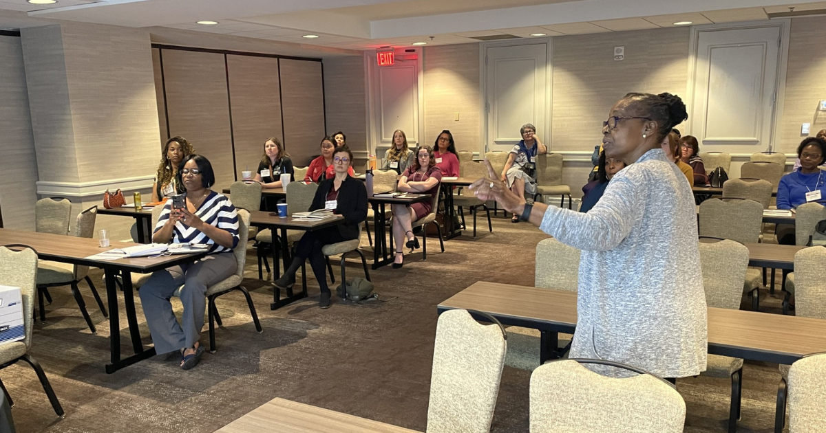 A woman stands and speaks to a room of people sitting at tables.
