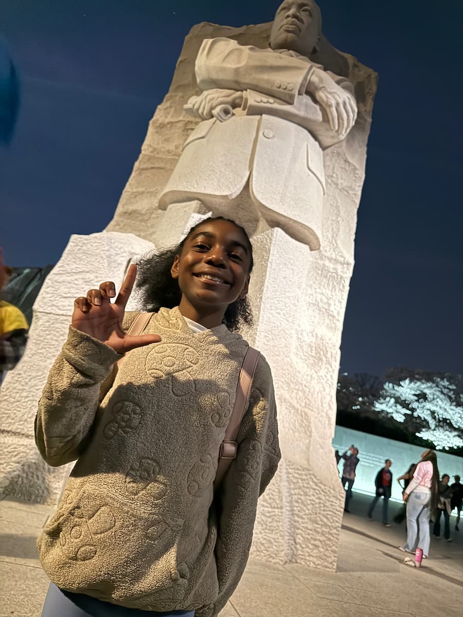A girl standing in front of a large stone statue of a man makes an “L” with her fingers.