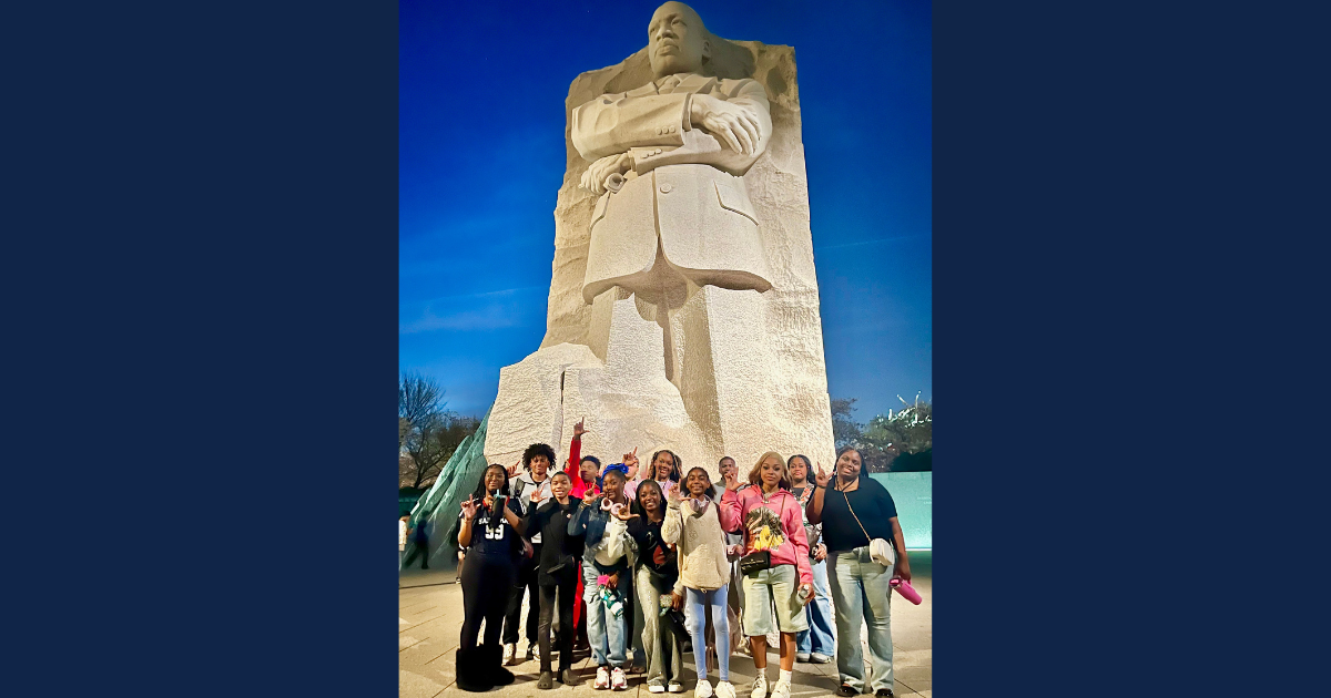 A group of youths make “L” shapes with their fingers in front of a large stone statue.