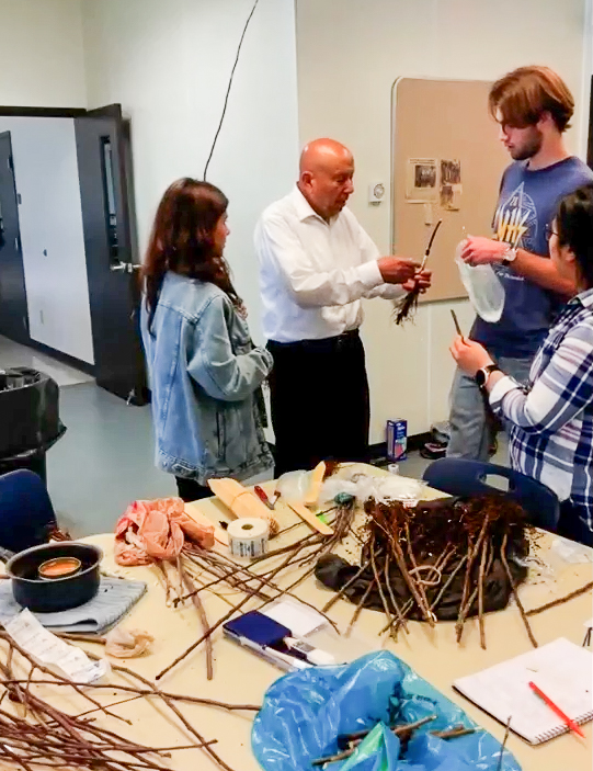 Four people standing around a table covered with various materials such as sticks, roots, leaves, tools, notebooks, and containers who are engaged in a hands-on workshop.
