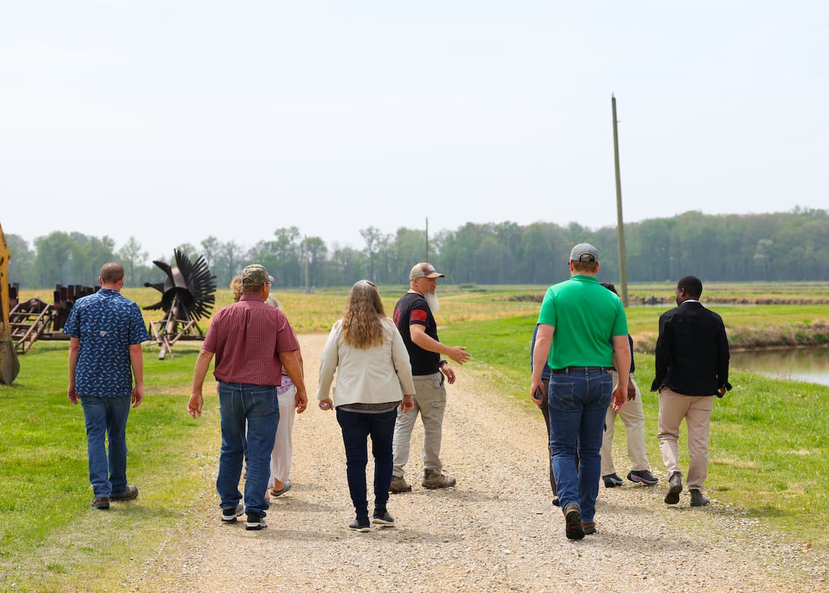 Group of people walk along a gravel road beside aquaculture ponds at NuTerra Farms while a man turns to speak with others.