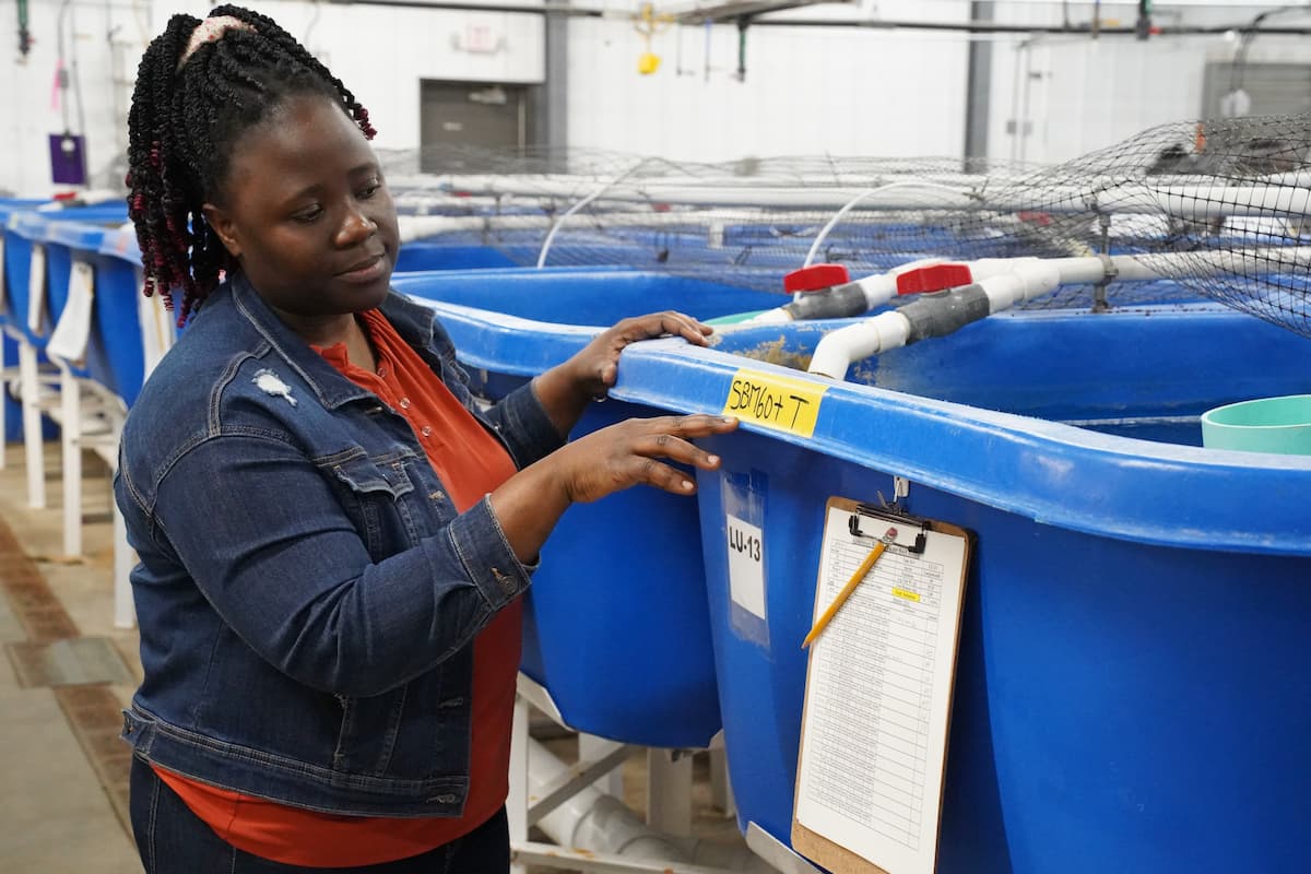 Dr. Moureen Matuha stands beside large blue research tanks at LU’s Aquaculture Research Facility and points to a tank label.