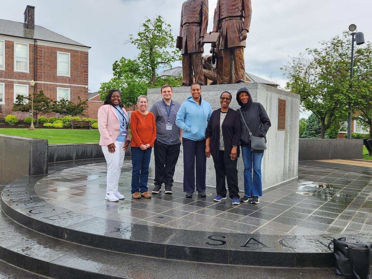 Six people standing outside in front of a large bronze statue on a circular stone platform with engraved words.