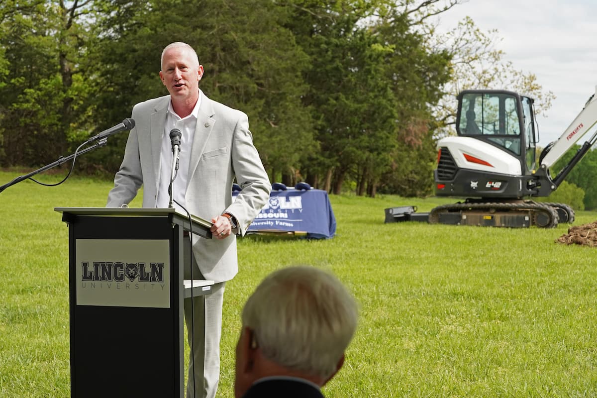 A man in a light gray suit speaking behind a Lincoln University podium outdoors on a grassy field. Background includes a blue Lincoln University table and a white excavator.