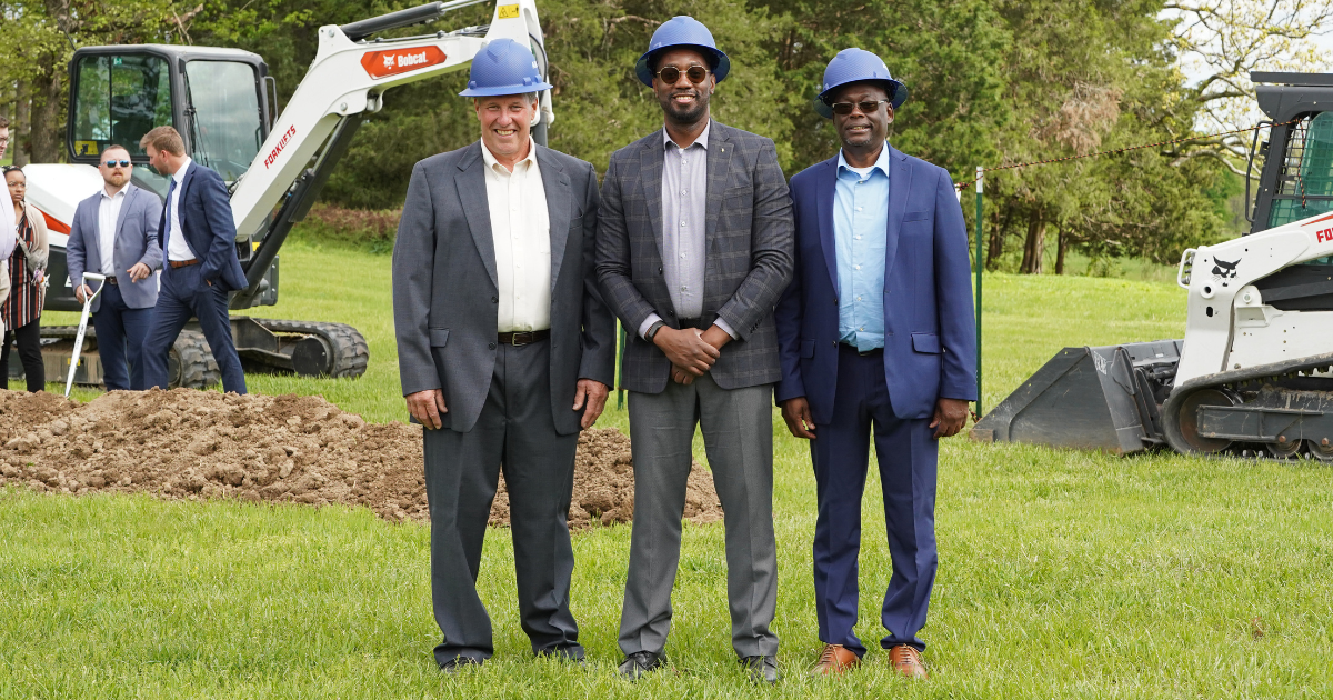 Three men wearing dark blue construction helmets and formal suits standing on grass in front of construction equipment and a dirt pile. 