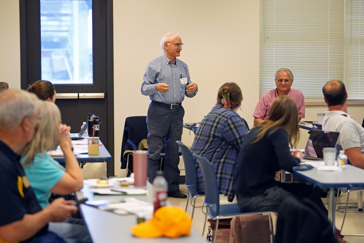 Many people sitting at tables watch and listen to a standing man speak.