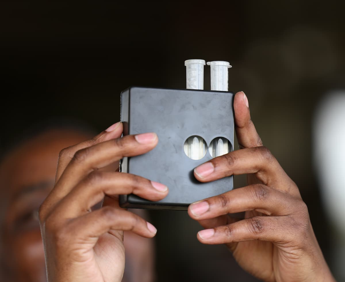 Close-up of Dr. John Maiyo’s hands holding a handheld water testing device with two sample tubes used to measure water quality.