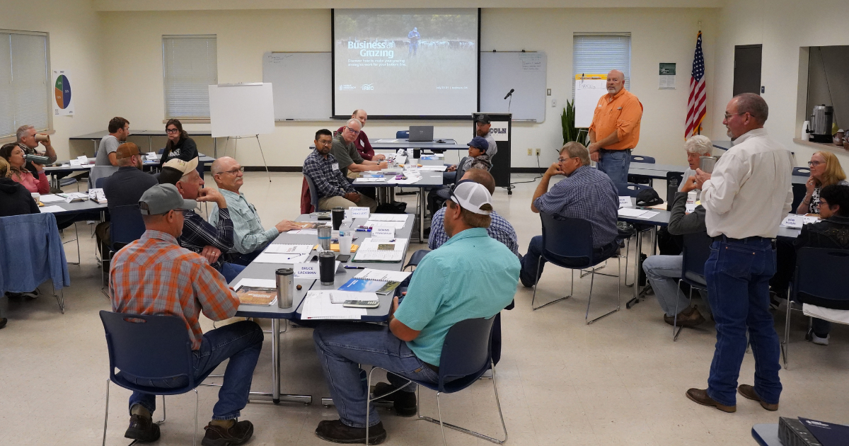 In a classroom setting, adults are seated around tables engaged in conversation. A presenter in an orange shirt stands near a projector screen displaying a slide titled "Business of Grazing."