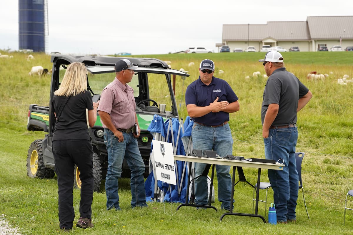 Four people standing around a folding table with a sign reading "Virtual Fencing" in a grassy outdoor area near a utility vehicle. 