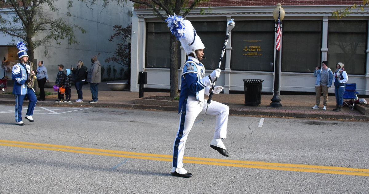 A drum major in a blue and white uniform marches down the street during a parade.