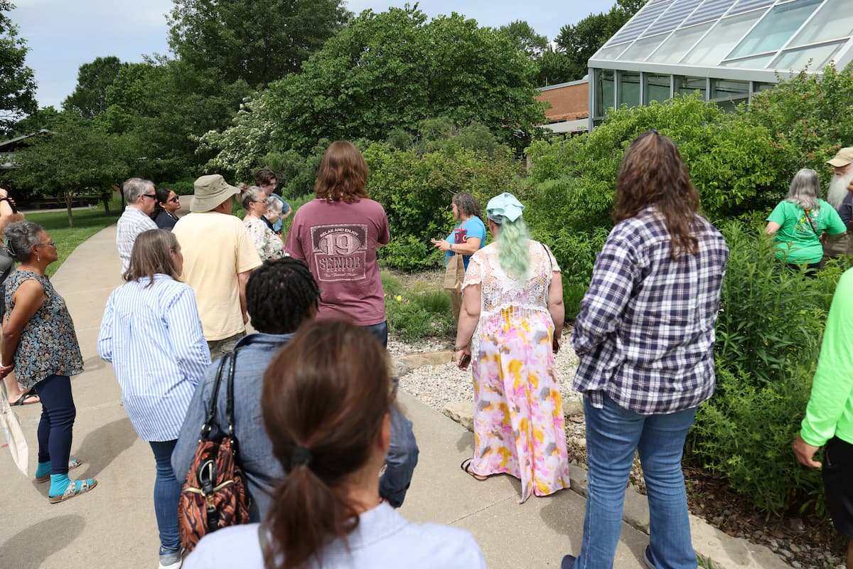 A group of people stands outdoors near garden plants and a greenhouse during a guided Native Plant Academy tour.
